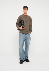 Brązowy sweter dzianinowy, jasnoniebieskie dżinsy w stylu dzwonów, czarna torba. Modelka stoi w zrelaksowanej postawie, mając na sobie czarne buty na grubej podeszwie.