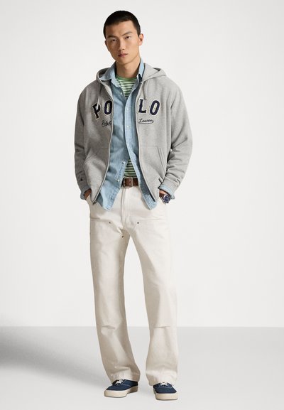 Άνδρας που φορά γκρι φούτερ με φερμουάρ και κείμενο "POLO", ανοιχτό μπλε τζιν πουκάμισο, πράσινη ριγέ μπλούζα, λευκό παντελόνι και μπλε παπούτσια, στέκεται με τα χέρια στις τσέπες.