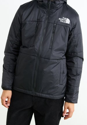 Mand iført en sort The North Face isoleret jakke med forreste lynlås og flere lynlåslommer, står foran en hvid baggrund.