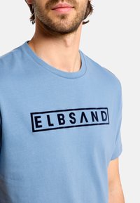 Svetlomodré bavlnené tričko s okrúhlym výstrihom; na hrudi má čierne obdĺžnikové logo s nápisom "ELBSAND" v tučnom písme.