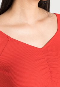 Top rosso a righe con scollatura a V, caratterizzato da una sottile texture e dettagli arricciati al centro. Inquadratura ravvicinata su scollatura e spalle.