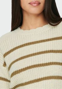 Pull en maille blanche présentant des rayures horizontales marron, un col côtelé et un tissu texturé avec une coupe légèrement ample.