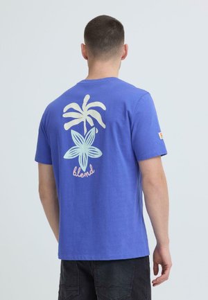 Uomo che indossa una maglietta blu con una grafica bianca di palma e fiore e la parola "blend" sul retro, abbinata a pantaloni neri.