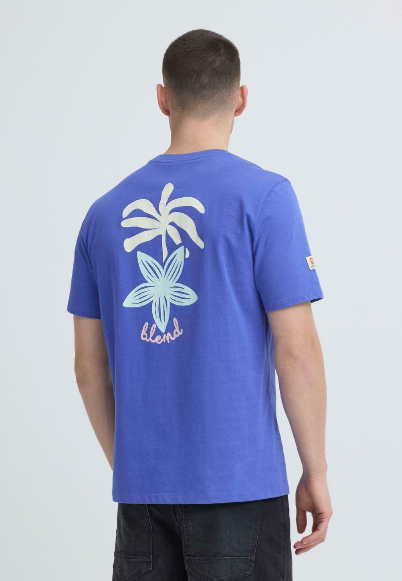 Άνδρας που φοράει μπλε μπλουζάκι με λευκό σχέδιο φοίνικα και λουλουδιού και τη λέξη "blend" στην πλάτη, συνδυασμένο με μαύρο παντελόνι.