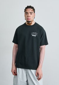 T-shirt en coton noir avec un col rond et des manches courtes, comportant un texte imprimé blanc "OBEY" et "movement" sur la zone gauche de la poitrine.