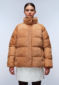 Brązowa kurtka puchowa z teksturą w prążki i wysokim kołnierzem. Nosi ją modelka na białej, pikowanej spódnicy z falistym wzorem.