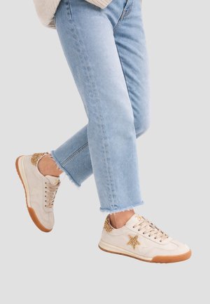 Persona che indossa jeans corti azzurri chiari e sneaker color crema con dettagli scintillanti dorati a forma di stella e sul tallone, in piedi davanti a uno sfondo semplice.