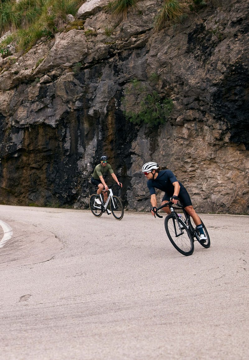 Deux cyclistes naviguent sur une route en asphalte courbée, portant des vêtements de sport ajustés et des casques. Un talus rocailleux se trouve en arrière-plan.