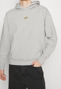 Sweat à capuche gris en mélange de coton, doté d'un petit logo jaune sur la poitrine et de poignets côtelés. Poche kangourou à l'avant incluse.