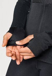 Zwarte sportjack met gestructureerde mouwen, duimhole en rits aan de voorkant. Glad stof met een aansluitend ontwerp en een slanke silhouet.