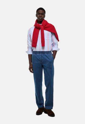 Jeune homme portant un pull rouge drapé sur les épaules, une chemise à rayures boutonnée, un jean à taille élastique bleu et des chaussures marron, debout les mains dans les poches.