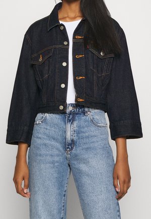 Femme portant une veste en jean foncé courte avec des surpiqûres orange sur une chemise blanche et un jean bleu clair taille haute.
