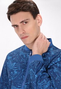 Giacca bomber blu con un motivo a foglie, colletto e polsini a costine, e chiusura a zip, realizzata in tessuto liscio con una leggera lucentezza.