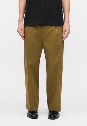 Άνδρας που φοράει φαρδιά, ελιάς πράσινα παντελόνια με μαύρα παπούτσια και μαύρο πουκάμισο, στέκεται μπροστά σε απλό λευκό φόντο.