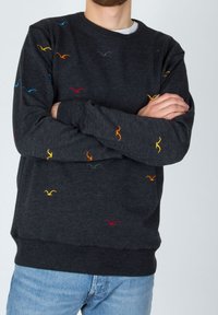 Uomo che indossa un maglione grigio scuro con motivi di uccelli ricamati colorati, braccia incrociate, abbinato a jeans azzurri chiari.