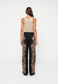 Woman with long hair wearing a sleeveless beige textured top and black wide-leg pants with pink and green floral embroidery, standing facing away.