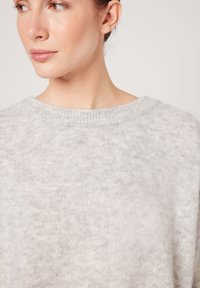 Naine, kes kannab helehallist, karvast pehmet villast kampsunit, millel on ümar kaelus, vaadates küljele ühevärvilise tausta ees.