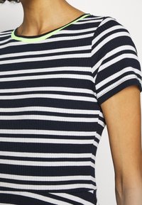 T-shirt côtelé à rayures bleu marine et blanc, manches courtes, avec un accent vert néon autour de l'encolure. Coupe ajustée avec une texture douce.