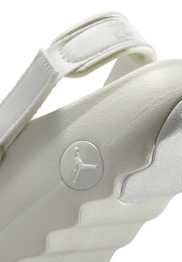 Biała sandał z teksturowaną gumową podeszwą, regulowanym paskiem oraz okrągłym detalem logo z wizerunkiem skaczącej postaci. Gładkie wykończenie i ergonomiczny kształt.