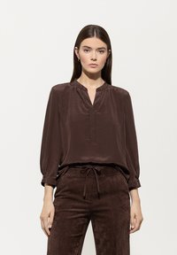 Braune Seidenbluse mit rundem Ausschnitt, knopflosem Frontverschluss und Dreiviertelärmeln. Getragen mit passenden strukturierten braunen Cordhosen.