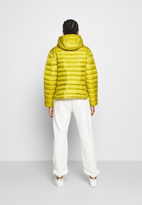 Keltainen puffytakki, jossa on vaakasuunnassa ommeltuja yksityiskohtia ja huppu, yhdistettynä valkoisiin verkkareihin ja tennareihin. Sujuva, kiiltävä pinta.