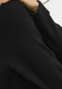 Czarny sweter o teksturowanym prążkowanym wzorze i luźnym kroju. Zbliżenie na rękaw, ukazujące miękki materiał.