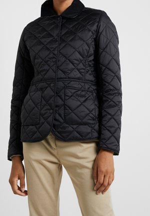 Femme portant une veste matelassée noire avec boutons-pression et deux poches avant, associée à un pantalon beige sur un fond uni.