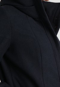 Cappotto di lana blu scuro con colletto alto e cuciture sartoriali, indossato da una persona che aggiusta il colletto con una mano alzata.