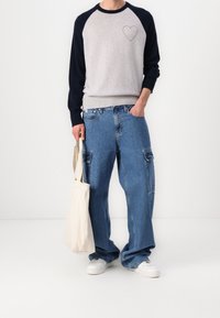 Personne portant un pull à manches longues gris et noir avec un motif de cœur, un jean cargo bleu ample, des baskets blanches, et tenant un sac fourre-tout beige.