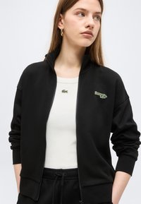 Νεαρή γυναίκα που φοράει μαύρο μπουφάν με φερμουάρ πάνω από ένα λευκό πουκάμισο με πράσινα λογότυπα κροκοδείλων, στέκεται μπροστά σε ένα απλό ανοιχτόχρωμο φόντο.