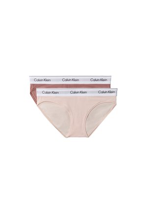 Femme debout pieds nus portant un bralette mauve Calvin Klein et un sous-vêtement assorti avec des bandes élastiques blanches siglées sur un fond gris.