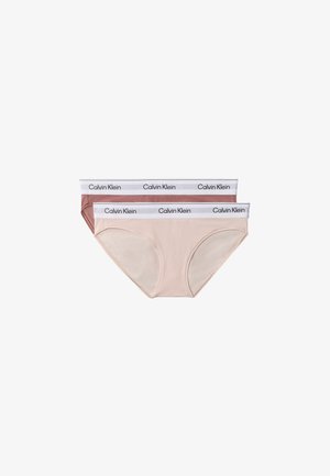 Femme debout pieds nus portant un bralette mauve Calvin Klein et un sous-vêtement assorti avec des bandes élastiques blanches siglées sur un fond gris.