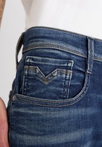 Gros plan sur un jean en denim bleu avec une petite poche avant présentant un design cousu en forme de V et une passants de ceinture visible.