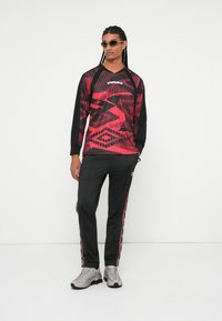 Langärmliges rotes und schwarzes Grafikoberteil mit geometrischen Mustern, kombiniert mit schwarzen Hosen mit rosa Seitenstreifen und grauen Sportschuhen.