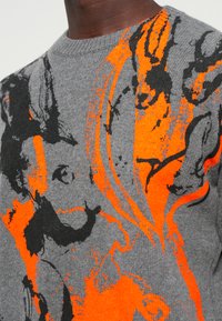 Grå sweater med abstrakte sorte og orange mønstre; rund halsudskæring; medium strikket tekstur. Tydelig visuel kontrast mellem farverne.