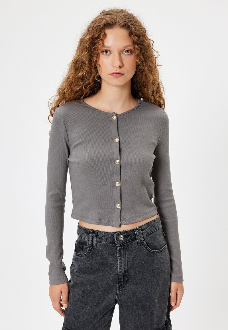 Grå, ribbet cardigan med lange ærmer i en cropped design og guldtonet knaplukning, båret over højtaljede sorte bukser.