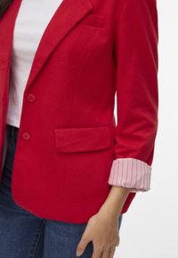 Blazer rouge à coupe ajustée, fermeture à un bouton et poche poitrine. Les manches retroussées présentent un motif rayé rose pâle et blanc.
