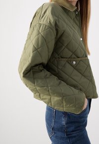 Quiltet olivengrøn jakke med krave, trykknapper og en frontlomme. Har diamantmønster og cropped design, lavet af polstret materiale.