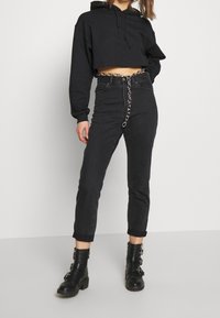 Giovane donna che indossa un hoodie nero cropped, jeans neri a vita alta con cintura a catena e stivali neri alla caviglia con fibbie, in piedi su sfondo bianco.