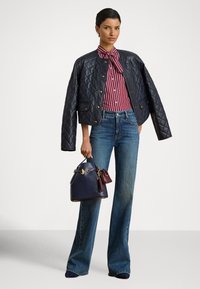 Μπλε σκούρο μπουφάν τύπου bomber με ρίγες πάνω από κόκκινο πουκάμισο με ρίγες, ανοιχτό μπλε τζιν καμπάνα, κρατώντας μια μπλε σκούρα τσάντα με κόκκινες λεπτομέρειες.