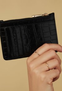 Porte-cartes en simili cuir noir avec un motif crocodile texturé, fermeture éclair et quincaillerie dorée. Main tenant le porte-cartes.