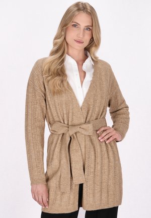Beige neulottu villatakki vyöllä vyötäröllä, jossa on ribbikuvio ja avoin etusuunta. Tyylitetty valkoisen paidan päälle.