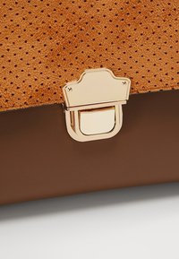 Brązowa skórzana torba z beżowym, perforowanym zamszowym klapem. Wyposażona w złoty metalowy zamek o k�ątowym kształcie, kontrastujące tekstury i kolory.