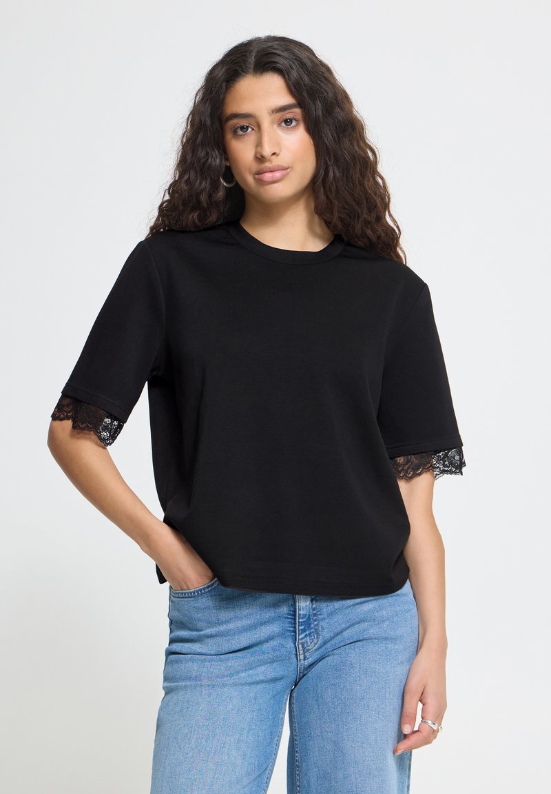 Femme aux longs cheveux bouclés portant un haut noir à manches courtes avec bordure en dentelle et un jean bleu clair, debout avec une main dans la poche.