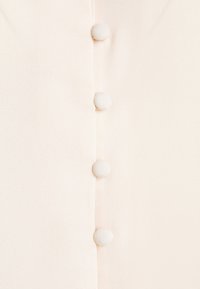Helle beige Bluse mit Knopfleiste, ausgestattet mit sechs stoffbezogenen Knöpfen und einer glatten, leicht strukturierten Oberfläche.