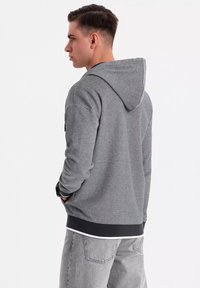 Sudadera gris de tela suave, con capucha ajustable, puños acanalados y borde negro. Lleva puesta unos vaqueros gris claro.