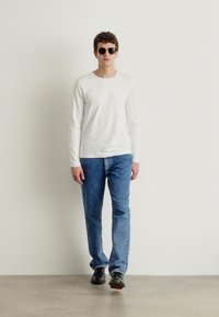 Hvit langermet skjorte, blå jeans og svarte sko. Har runde svarte solbriller. Minimalistisk design, nøytrale farger og avslappet passform.