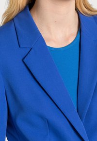 Blazer bleu avec un col cranté, confectionné dans un tissu lisse. Porté par-dessus un haut bleu côtelé. Coupe ajustée mettant en valeur un design structuré.