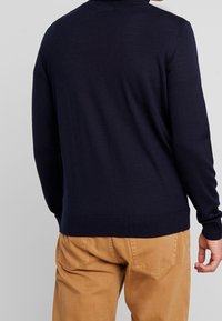 Mörkblå stickad tröja med rund halsringning och långa ärmar. Materialet verkar mjukt med ribbade muddar och fåll. Bärs med bruna byxor.