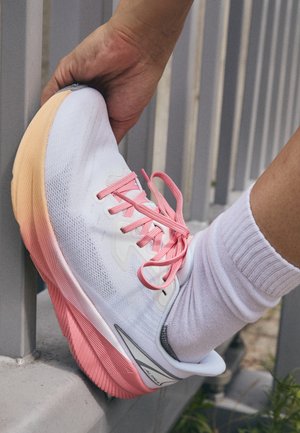 White athletic shoe with pink laces, textured mesh upper, and a gradient pink to orange sole. Sock visible above the ankle.